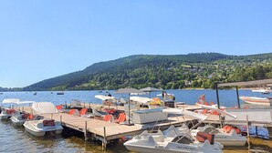 A marina with several boats docked, a wooden pier, and a backdrop of a forested hill with houses. - Lovely apartment in Gérardmer with house sea view (Gérardmer)