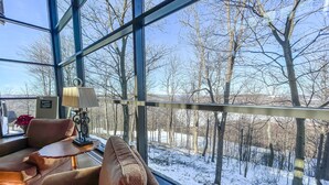A room with a large window overlooking a snowy landscape, a sofa with cushions, a lamp, and a small table.