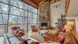 A spacious living room with leather sofas, a stone fireplace, large windows, and wooden beams.