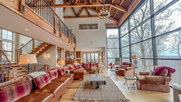 A spacious living area with a high ceiling, wooden beams, a large window, and a staircase leading to an upper level.