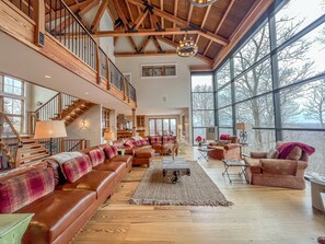 A spacious living area with a high ceiling, wooden beams, a large window, and a staircase leading to an upper level. - Stunning Luxury Home! Direct Ski Access and Hot Tub (McHenry)