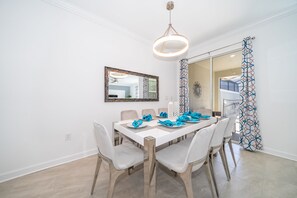 Un comedor con una mesa blanca dispuesta para seis personas, rodeada de sillas. Un espejo grande cuelga de la pared, y se ve parcialmente una cortina estampada.