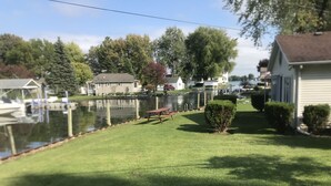A residential area with a grassy lawn, a small bush, and a dock with boats. - Cheerful 3 bedroom cottage with River access. (Clay Township)