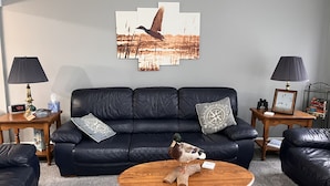 A living room with a black leather sofa, two wooden side tables with lamps, a coffee table, and a wall-mounted artwork of a bird in flight. - Cheerful 3 bedroom cottage with River access. (Clay Township)