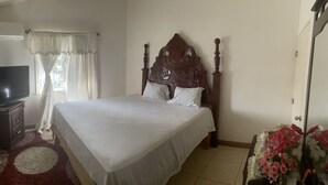 A neatly made bed with a wooden headboard, a television on a stand, and a floral arrangement on a side table. - Spacious 9 BED family sized house in a gated community (Trelawny Parish)