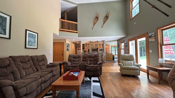 A spacious living room with a high ceiling, a wooden ceiling fan, and a view of the kitchen and dining area through an open door.