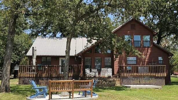 A red wooden cabin with a porch, surrounded by trees and a grassy area with a bench and chairs. - A Dream Come True Lake House  - The prettiest sunset view on the lake! (Marquez)