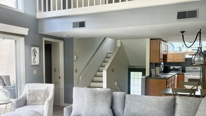 Ein geräumiges Wohnzimmer mit einem großen grauen Ecksofa, einem Holztisch und einem weißen Sessel. Eine Treppe führt zu einer oberen Ebene mit einem Balkon.