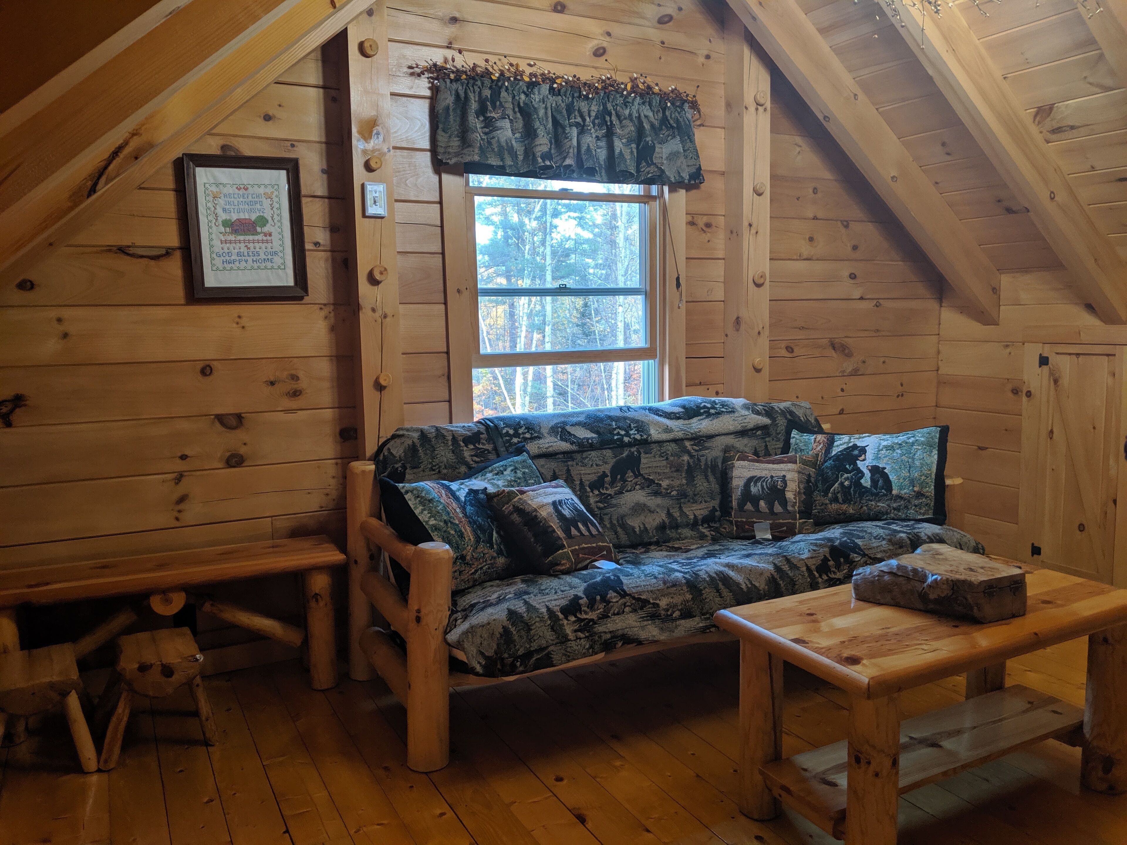 A cozy cabin with a wooden interior, a sofa with bear-themed cushions, a small wooden table, and a window with a view of trees.