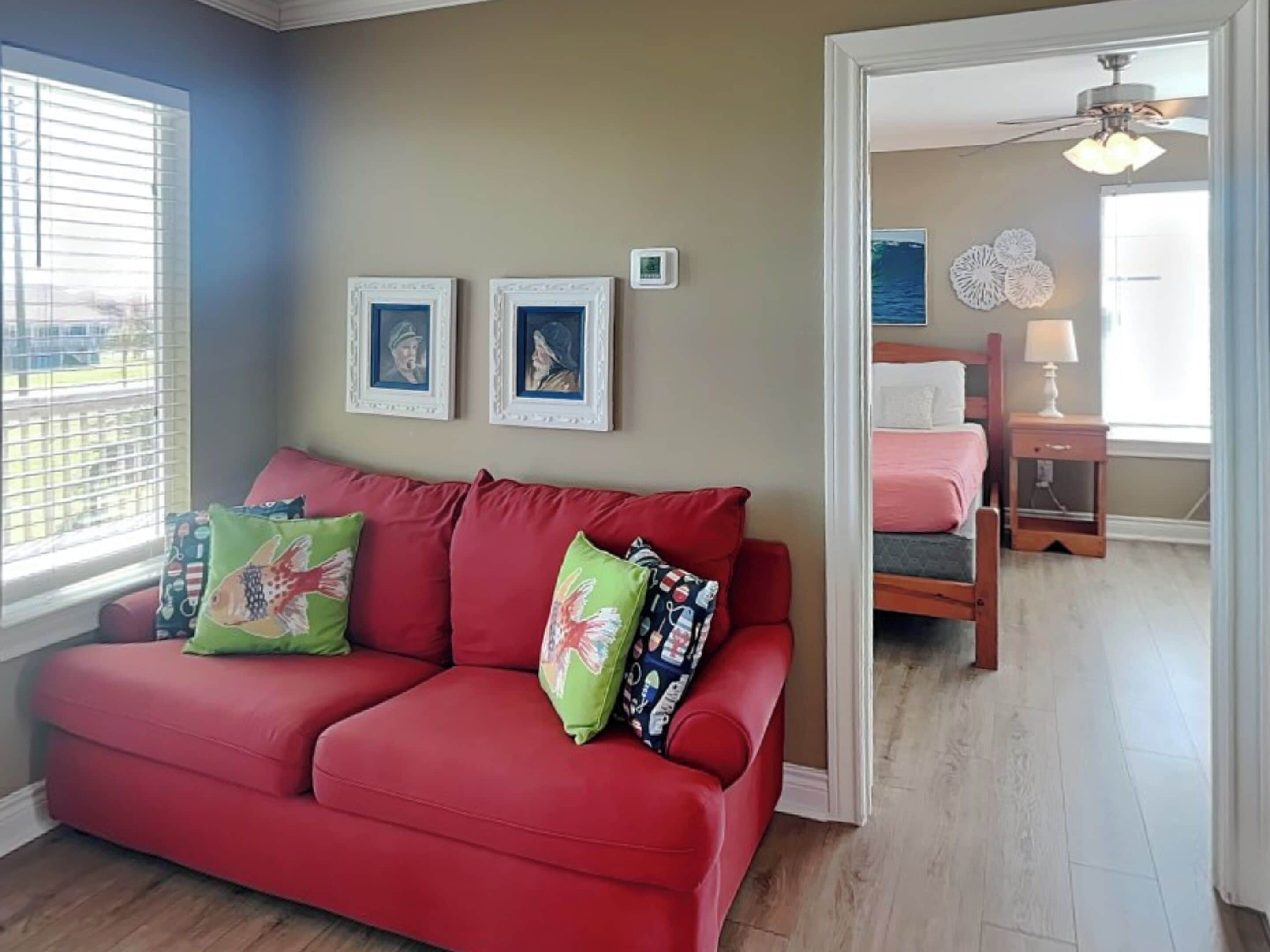 A red sofa with patterned pillows, a ceiling fan, a bed with a pink blanket, and framed pictures on the wall.