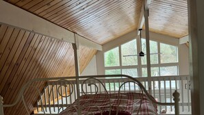 Un dormitorio con techo inclinado, cama con estructura metálica y ventanas amplias que dejan entrar luz natural.