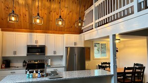 Cocina moderna con armarios blancos, encimeras de granito y electrodomésticos de acero inoxidable. Se observan tres lámparas colgantes, una isla grande con taburetes y un área de comedor con mesa y sillas.