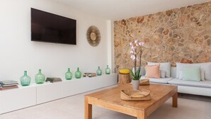 A modern living room with a stone wall, a flat-screen TV mounted above a white console, and a wooden coffee table with a woven basket. - CAN FONT (COSTITX) - Stunning villa with private pool and free WiFi. (Costitx)