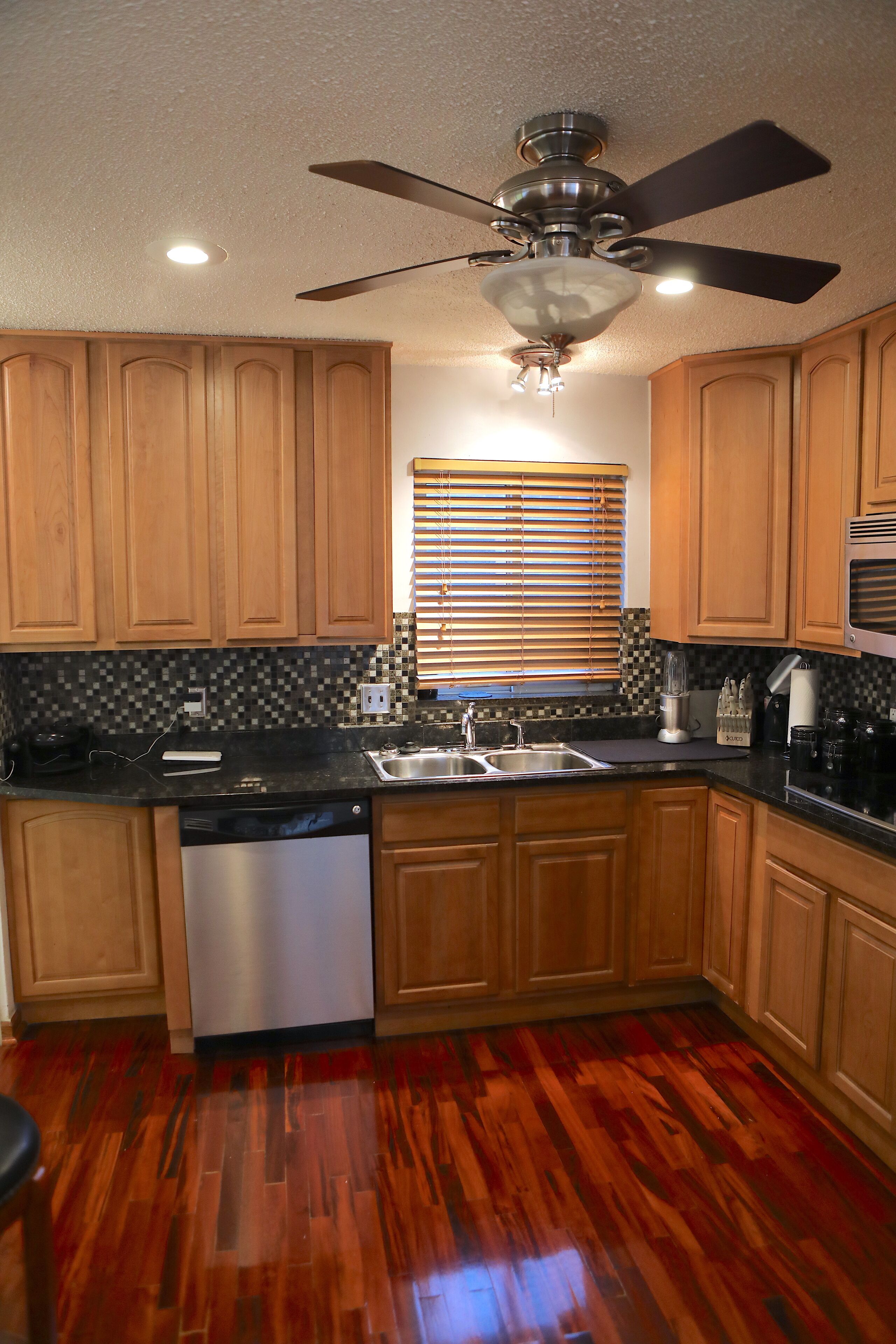 A kitchen with wooden cabinets, a black countertop, a dishwasher, a ceiling fan, and a window with blinds.