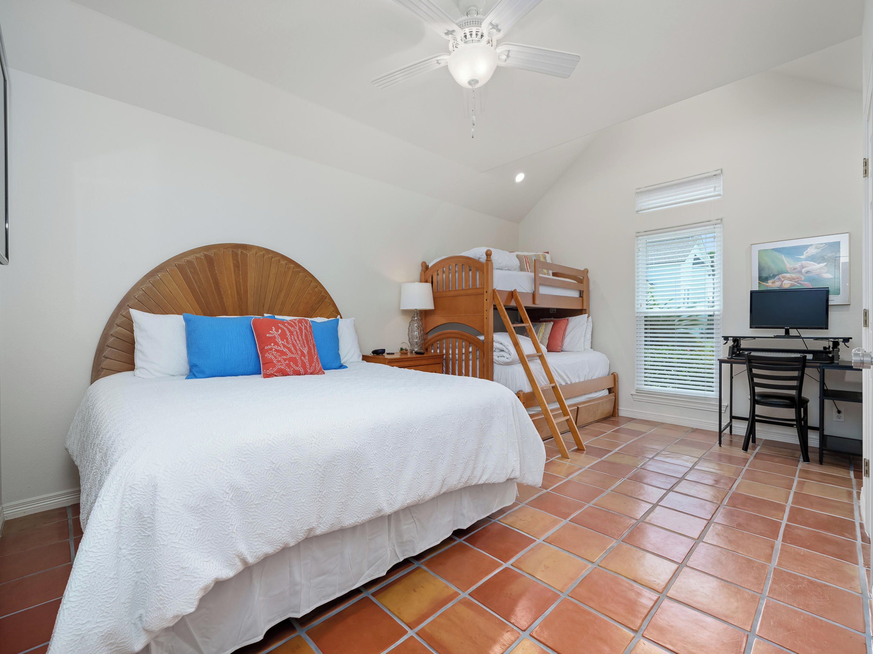 A bedroom with a large bed, a bunk bed, a desk with a chair, a ceiling fan, and a window with blinds.
