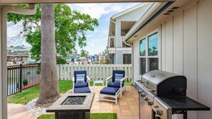 An outdoor patio with a grill, two chairs, a fire pit, and a view of a residential area. - Flamingo House by Miss Kitty’s (Rockport)