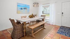 A dining area with a wooden table, wicker chairs, and a bench. There is a painting of boats on the wall, a hanging light fixture, and a door with a keypad lock. - Flamingo House by Miss Kitty’s (Rockport)