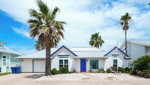 A single-story house with a blue door and white siding, surrounded by palm trees and a clear sky. - Flamingo House by Miss Kitty’s (Rockport)