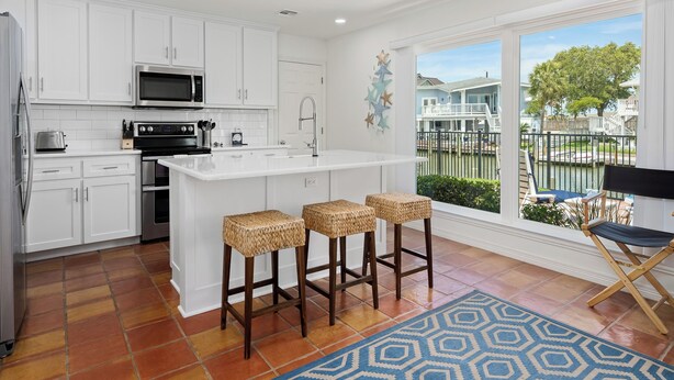 A modern kitchen with white cabinets, a large island with bar stools, a view of a residential area through large windows, and a blue patterned rug. - Flamingo House by Miss Kitty’s (Rockport)