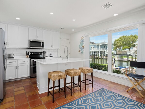 Una cocina moderna con armarios blancos, una isla grande con taburetes y vistas a una zona residencial a través de amplios ventanales, además de un tapete azul con estampado.