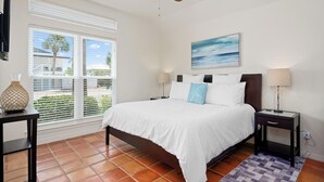 A bedroom with a bed, a ceiling fan, a window with blinds, a painting, and a nightstand with a lamp. - Flamingo House by Miss Kitty’s (Rockport)