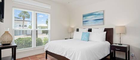A bedroom with a bed, a ceiling fan, a window with blinds, a painting, and a nightstand with a lamp.