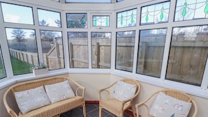 A corner room with wicker furniture, large windows, and a view of a wooden fence. - St Marys Cottages No1 (Alnwick)
