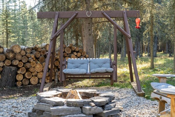 A wooden swing with a cushioned seat, a fire pit, and a stack of logs in a forest setting.