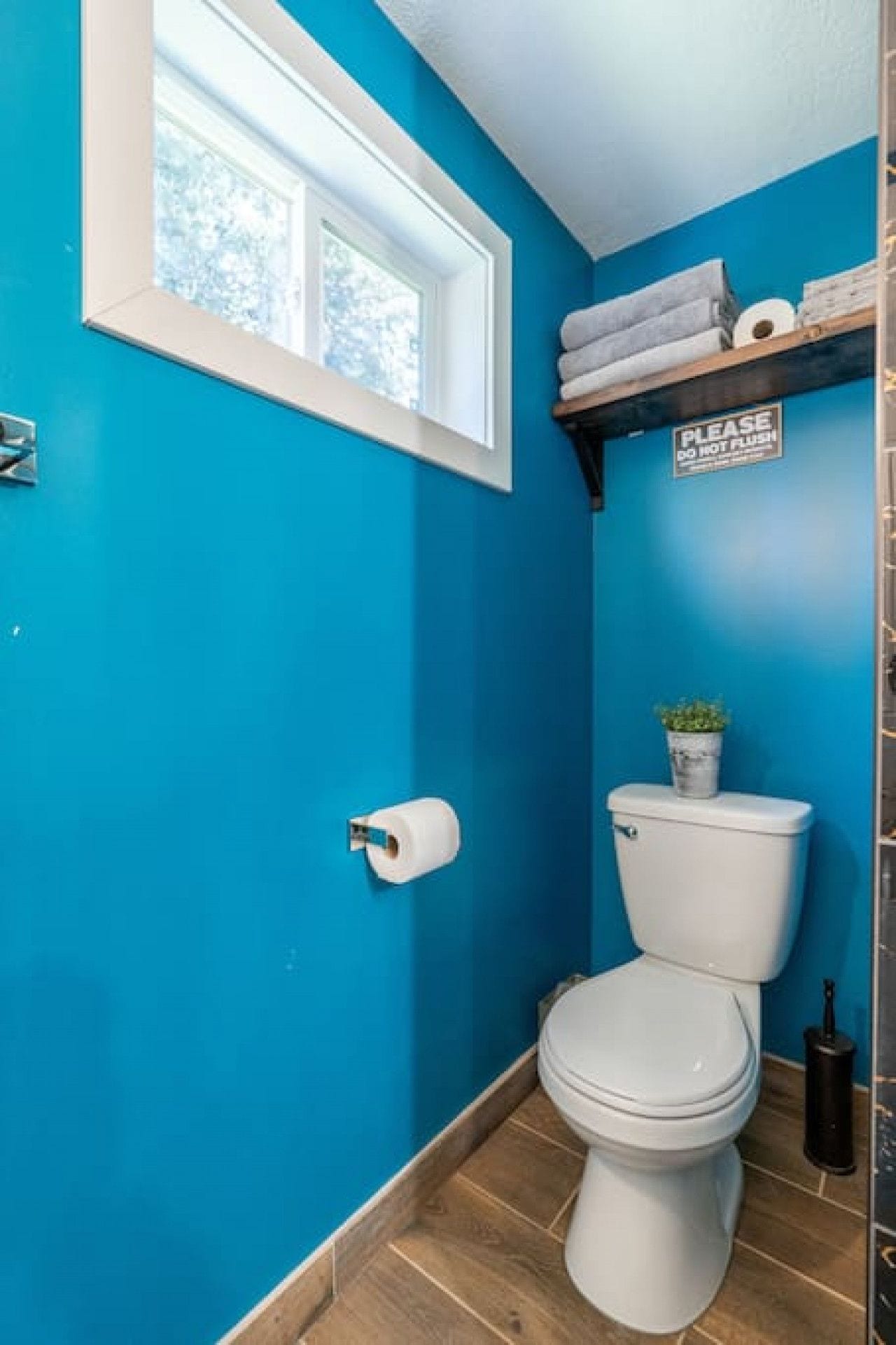 Une petite salle de bain avec des toilettes, un rouleau de papier toilette, une fenêtre et un panneau accroché au mur.
