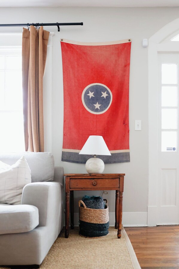 A living room with a sofa, a lamp on a wooden side table, a red flag with a circular emblem, and a window with beige curtains. - The Bellmore Cottage (Nashville)