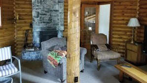 Interior de una cabaña de troncos con chimenea de piedra, dos sillones tapizados, una silla a rayas, una mesa de madera y una lámpara.