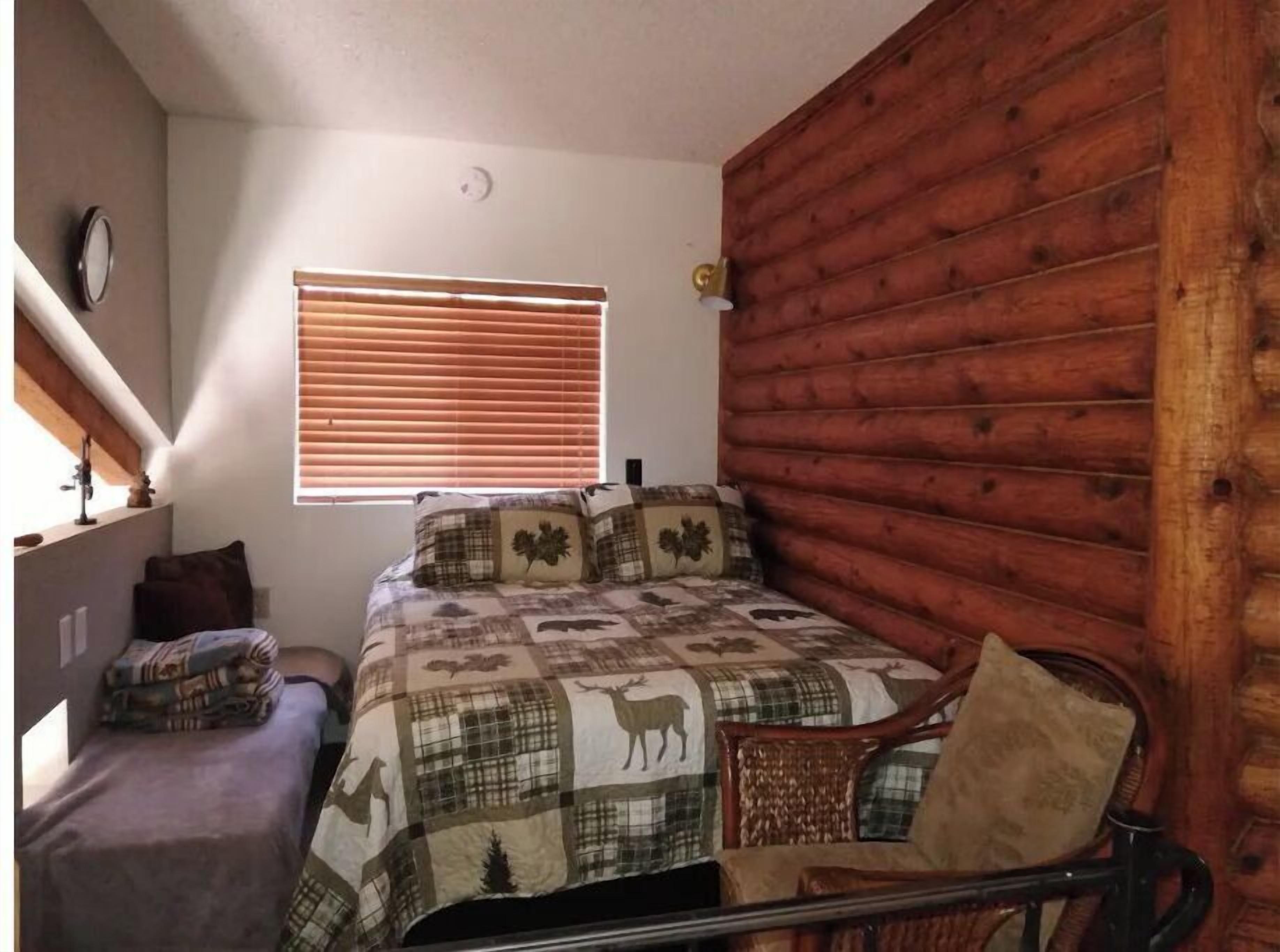 A bedroom with a bed, a wooden wall, a window with blinds, a chair, and a small table.