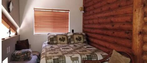 A bedroom with a bed, a wooden wall, a window with blinds, a chair, and a small table.