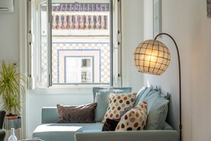 A room with a window, a sofa with patterned cushions, a floor lamp, and a potted plant.