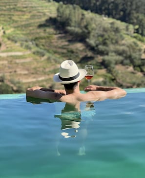 A person relaxing in a clear pool with a drink, surrounded by greenery and hills.