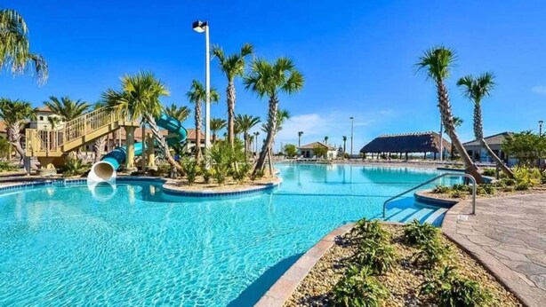 A swimming pool with a water slide, palm trees, and a covered area.