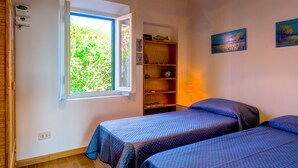 A bedroom with two beds, a window with a view of greenery, a shelf with decorative items, and wall art.