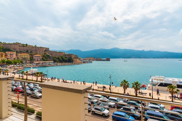A coastal cityscape with a marina, parked cars, and a view of the sea and mountains.