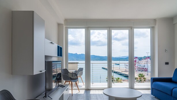 Een moderne woonkamer met een televisie, zitmeubels en een balkon met uitzicht op zee en bergen.
