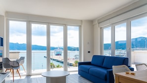 Een moderne woonkamer met een blauwe bank, een ronde tafel, en een balkon met grote ramen met uitzicht op een jachthaven en bergen.