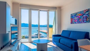 Ein modernes Wohnzimmer mit einem blauen Sofa, einem kleinen runden Tisch und einem Fernseher auf einem Schrank. Große Glasschübe führen zu einem Balkon mit Blick auf ein Gewässer und Boote.