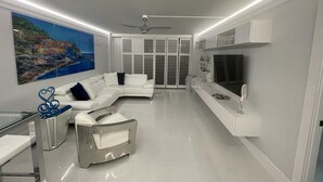 A modern living room with a white sectional sofa, a glass coffee table, a flat-screen TV mounted on the wall, and a ceiling fan. - Bay view 1st floor condo with beach access. (Marco Island)