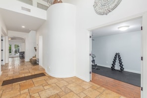 A spacious hallway with a curved white wall, a wooden floor, and a door leading to a room with exercise equipment. - 5BR Mesa Home 4600 sq.ft | Pool, Hot Tub, Putting Green, In-Ground Trampoline, Half Court Basketball (Mesa)