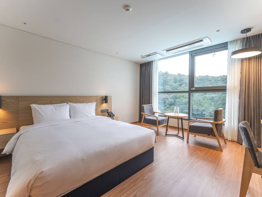 A modern hotel room with a large bed, a desk, two chairs, and a view of the outdoors.