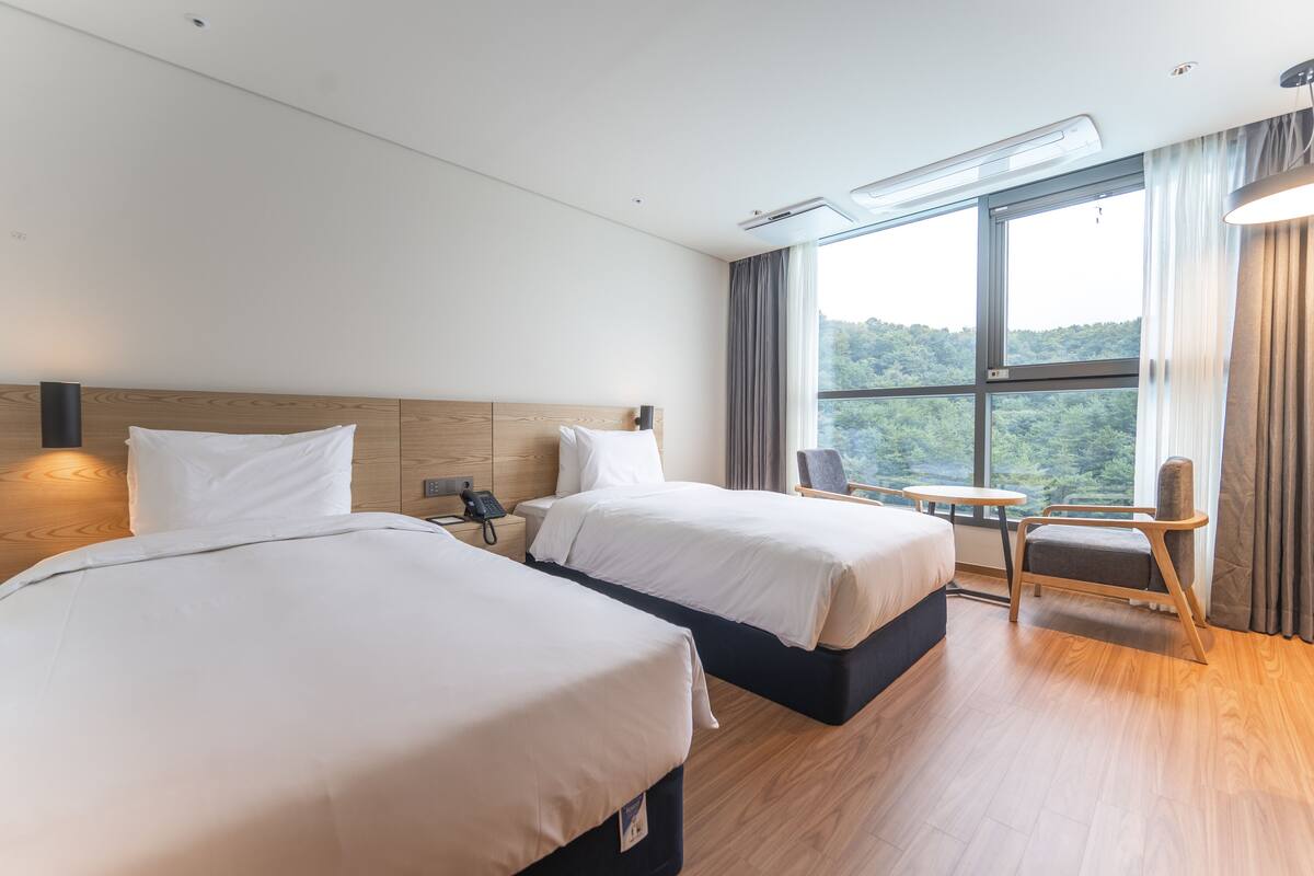 A hotel room with two beds, a small table, a chair, and a large window offering a view of the forest.