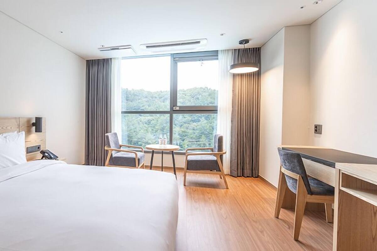 A hotel room with a large bed, a desk, two chairs, a small table, and a window with curtains.