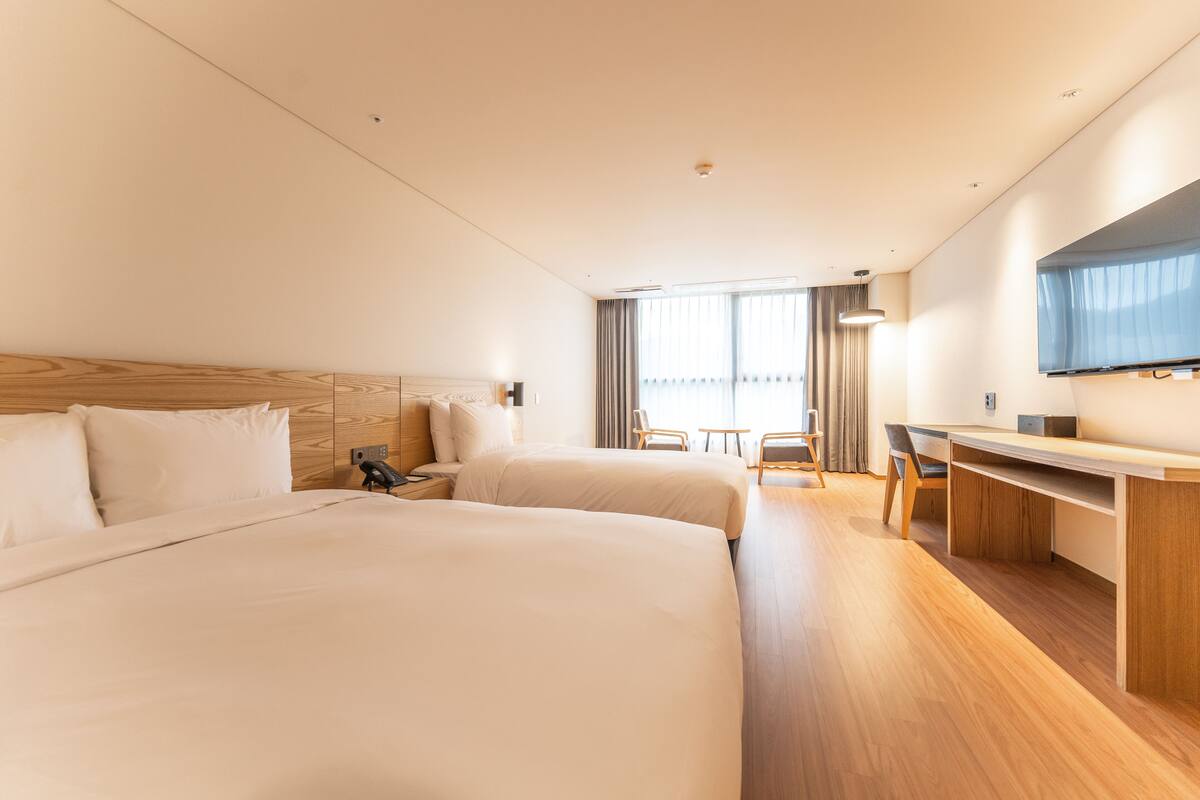 A hotel room with two beds, a desk, a chair, a TV, and a window with curtains.
