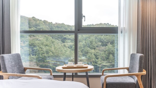A hotel room with a large window, two armchairs, a small round table with a tray of cups and saucers, and a bed with a white duvet. - Grand Moment (Busan)