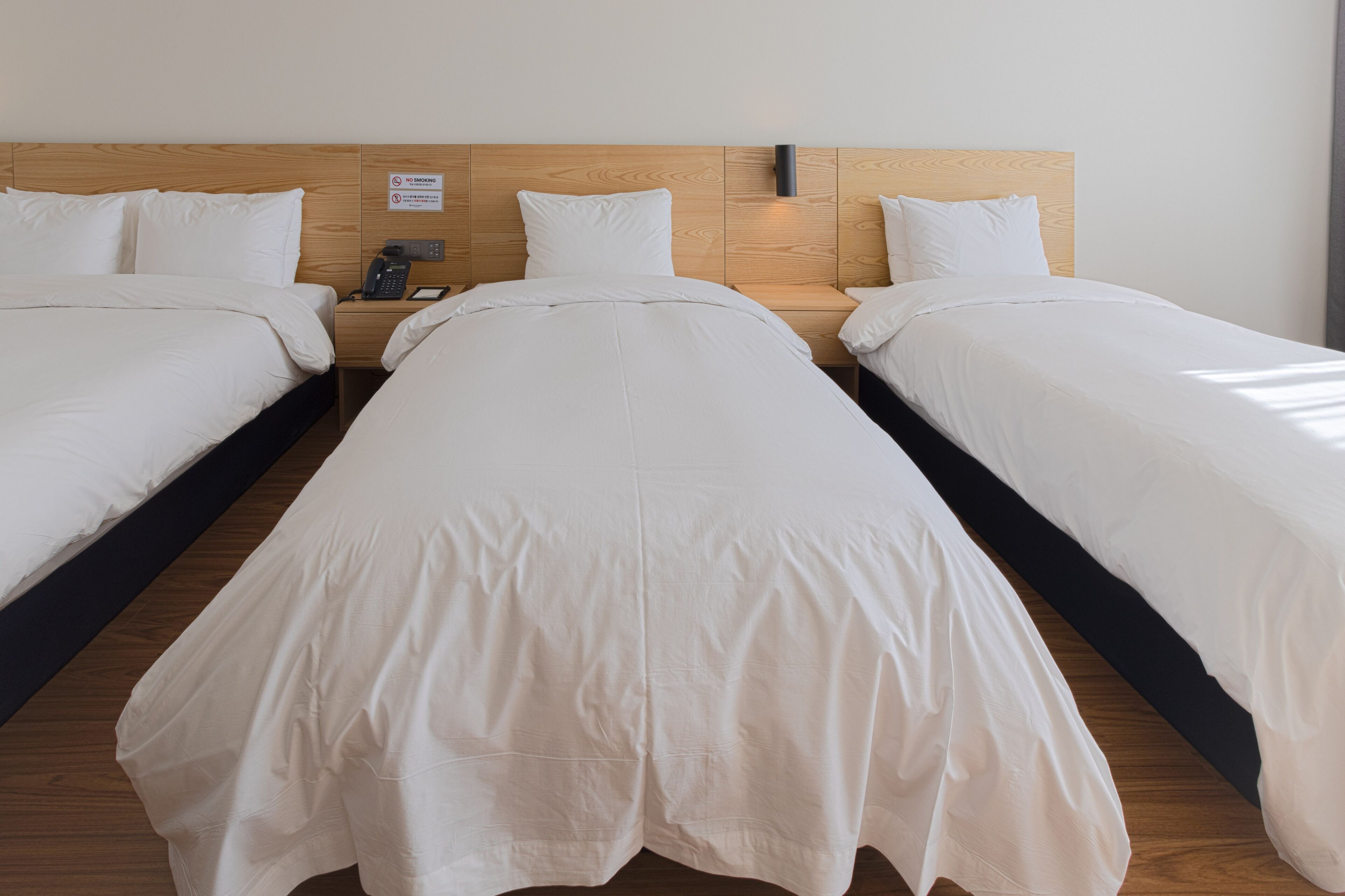 Camera d'albergo con tre letti singoli, una testiera in legno, un comodino con il telefono e un cartello che indica il divieto di fumare.
