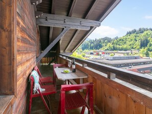 A wooden balcony with a table and chairs, overlooking a forested area with buildings in the distance.